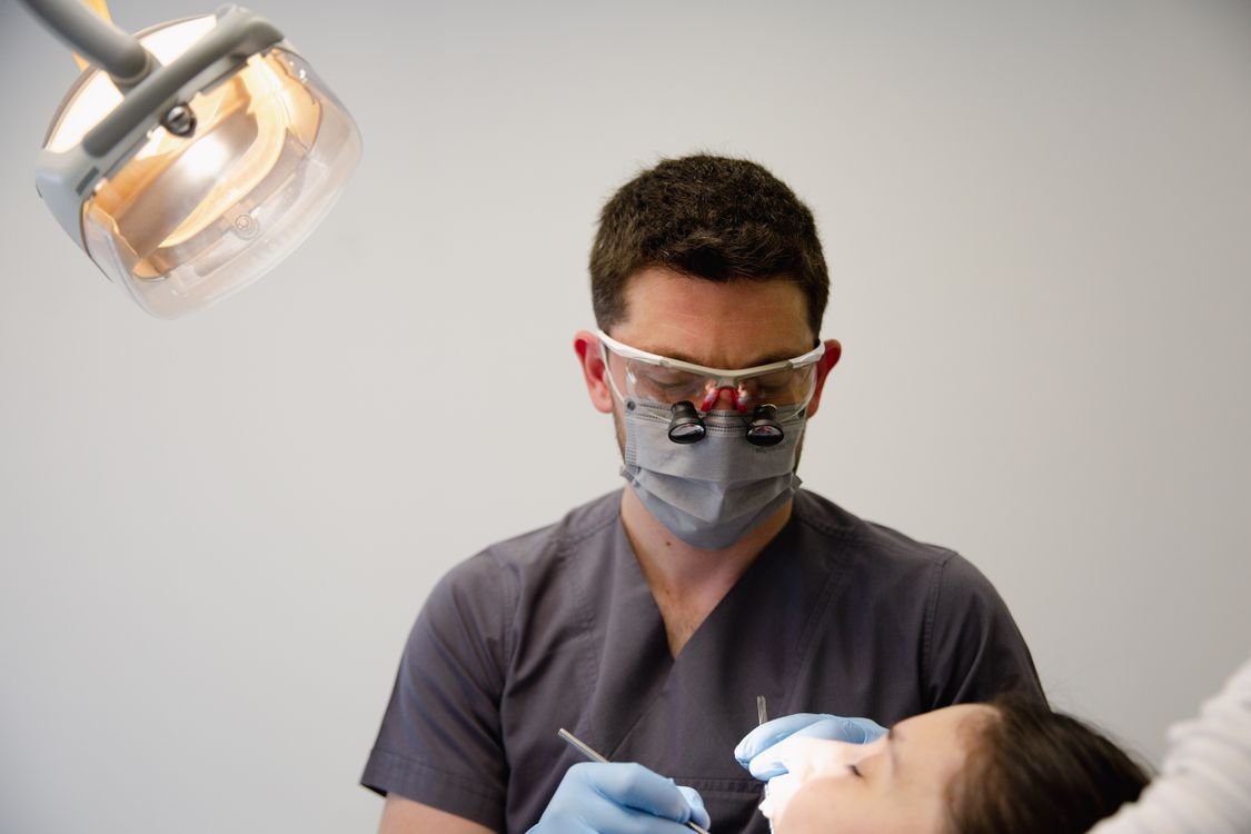 A masked dentist wearing surgical loupes performs a procedure on a patient under an overhead operatory light.