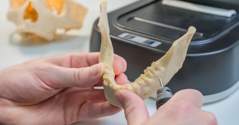 Gloved hands holding a 3D-printed dental model in a digital dental lab, with a 3D printer visible in the background.