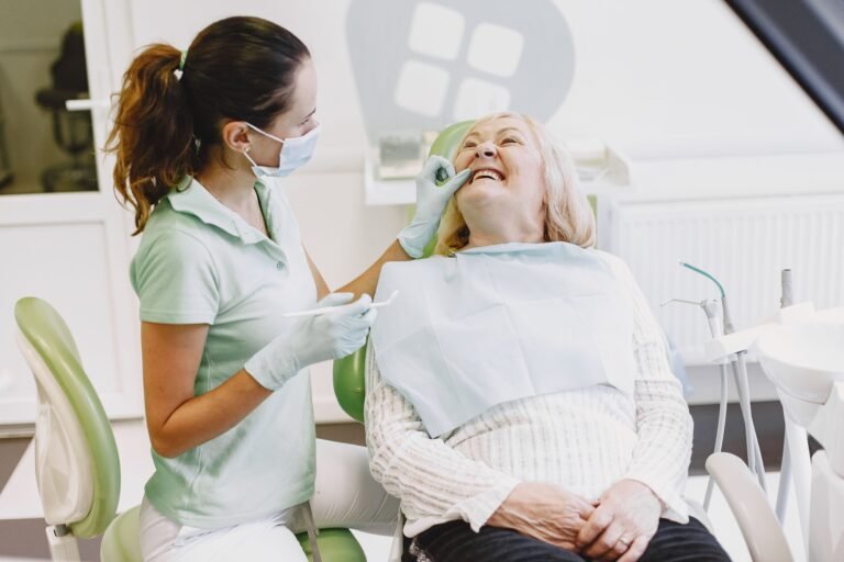 A female dentist examines a senior woman patient in a dental office, both smiling during the appointment.