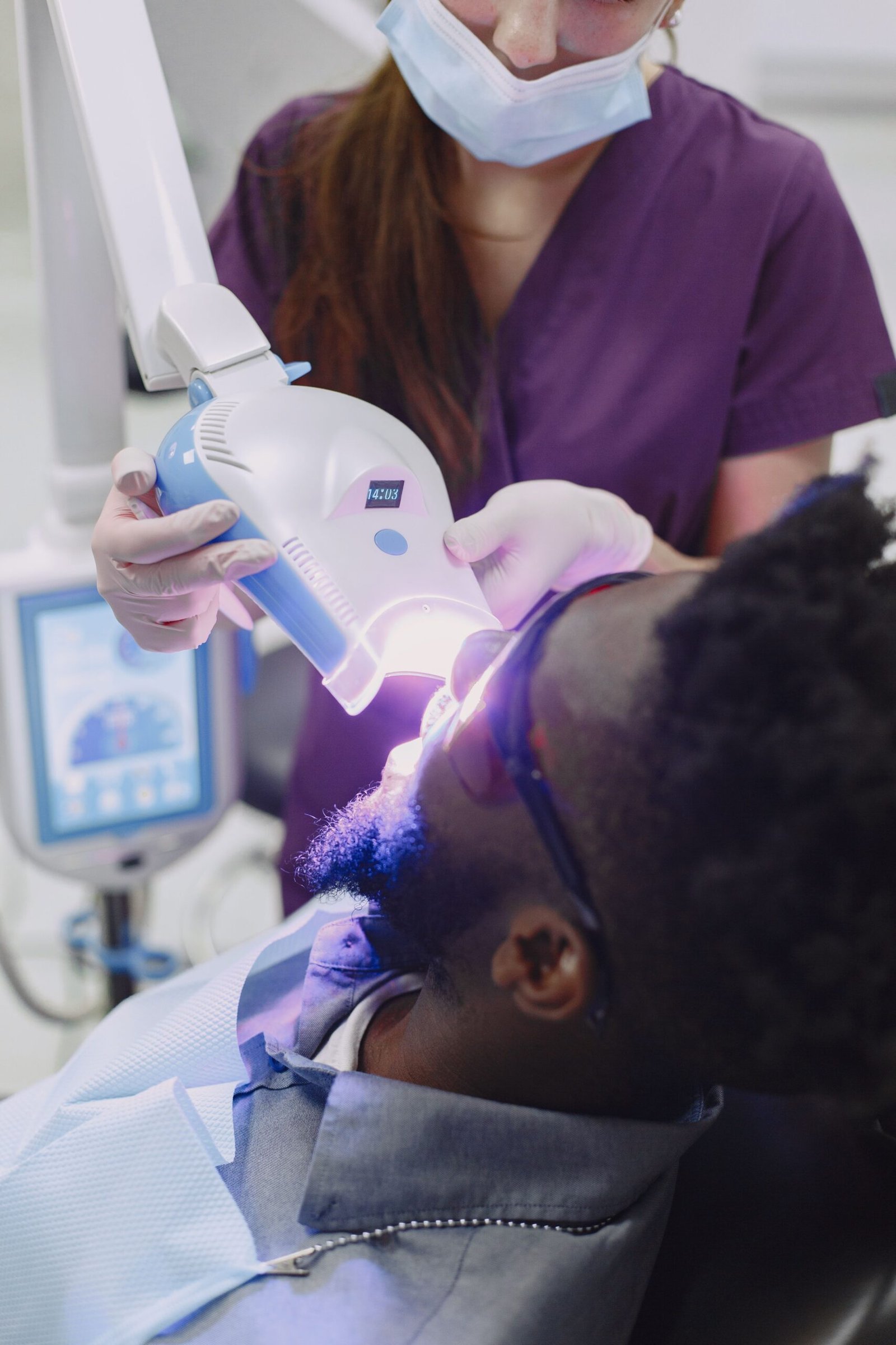 A dental professional directing a handheld light at a patient's teeth during a whitening procedure.