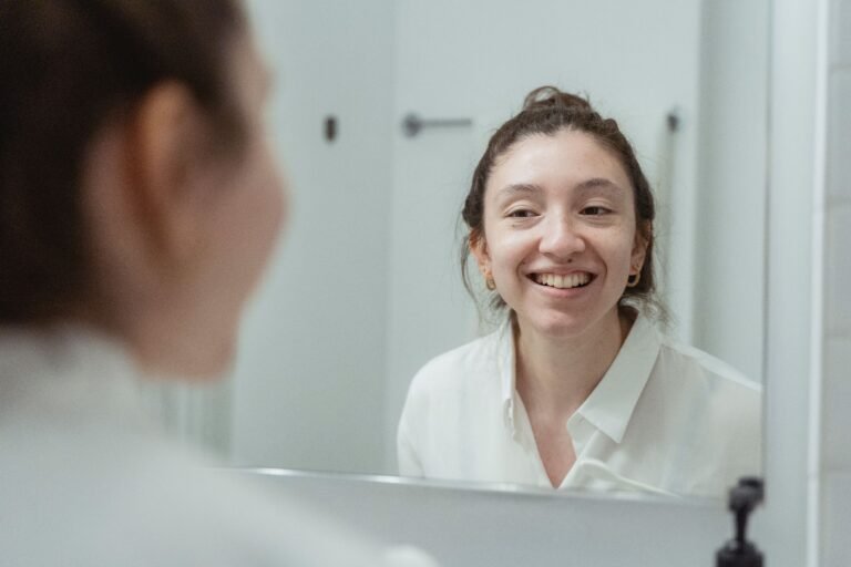 A woman in a white shirt smiling at her reflection in a bathroom mirror.