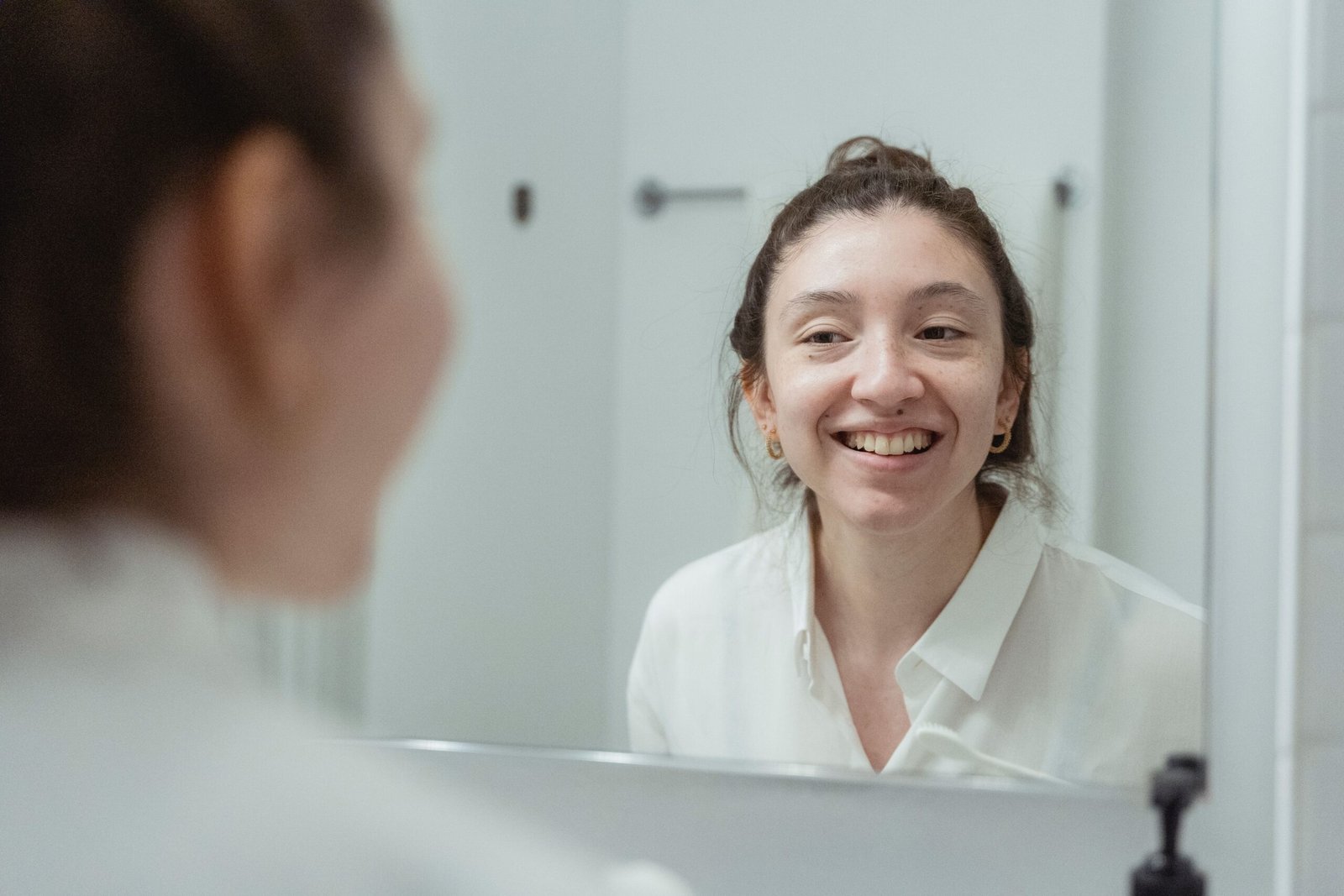 A woman in a white shirt smiling at her reflection in a bathroom mirror.